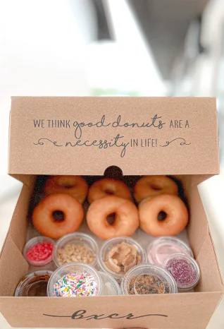 Natural brown kraft four corner donut kit box with bold black typography on the inside lid reading “We think good donuts are a necessity in life!” and subtle branding “boxr” along the bottom edge.
