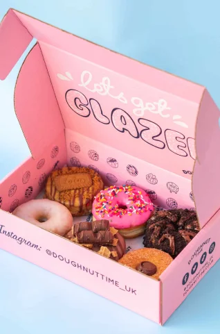 Bright pink Doughnut Time UK box with playful inside lid text “let’s get GLAZED!” and branded side print including Instagram handle. Inside: assorted decorated doughnuts.