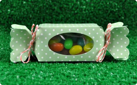 Custom-printed candy-shaped box with matte pastel green polka dot design, die-cut oval window, and twisted ends tied with pink and white thread rope.